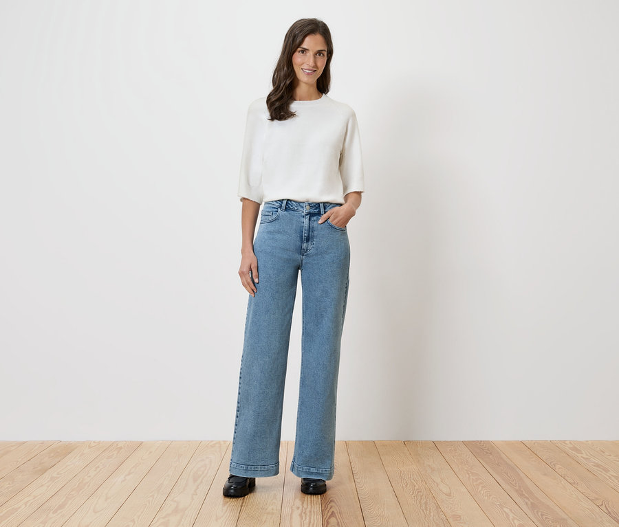 Žena stojí pred bielou stenou a má na sebe bielu blúzku a široké džínsy.