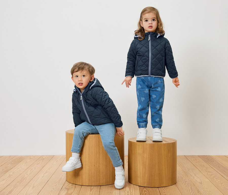 Chlapec sedí na drevenej stoličke, zatiaľ čo dievča stojí na inej. Obaja majú na sebe tmavomodré prešívané bundy a džínsy.