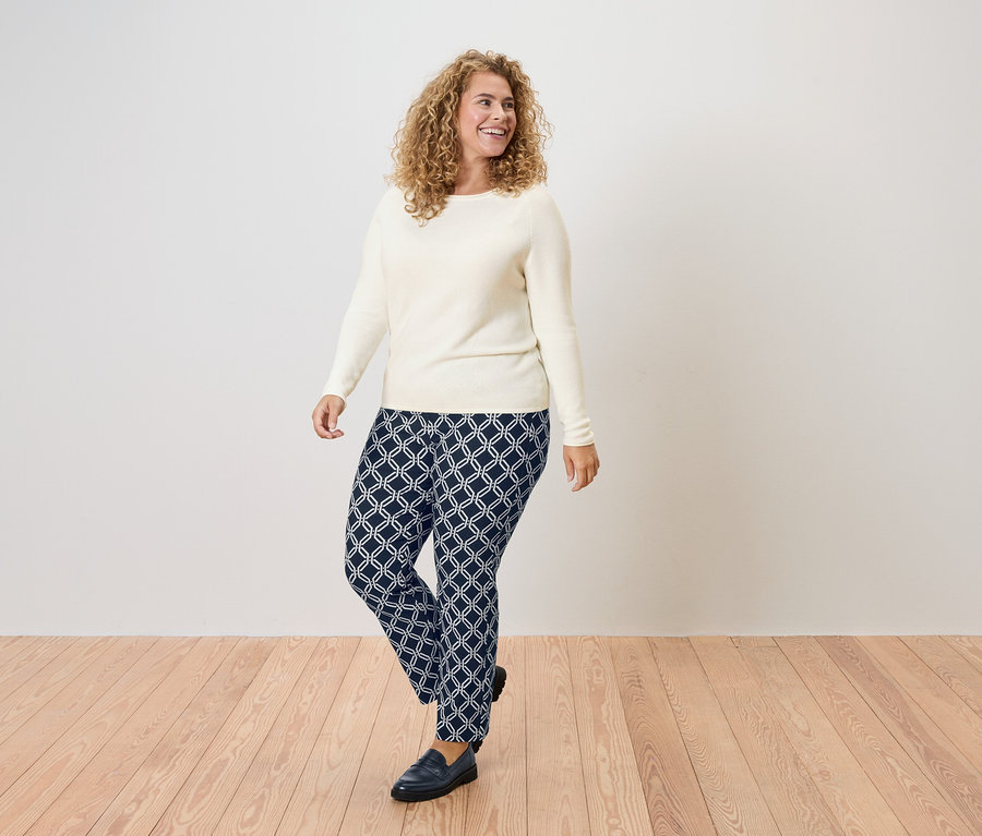 Žena s kučeravými vlasmi v krémovom svetri a tmavomodrých nohaviciach s geometrickým vzorom.