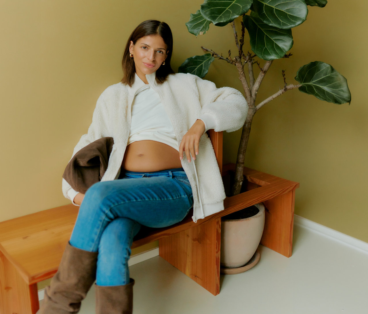 Tehotná žena sedí na lavičke vedľa rastliny a má na sebe otvorenú plyšovú bundu MAMALICIOUS 3 v 1 »MLCARLY« cez biele tričko a džínsy.
