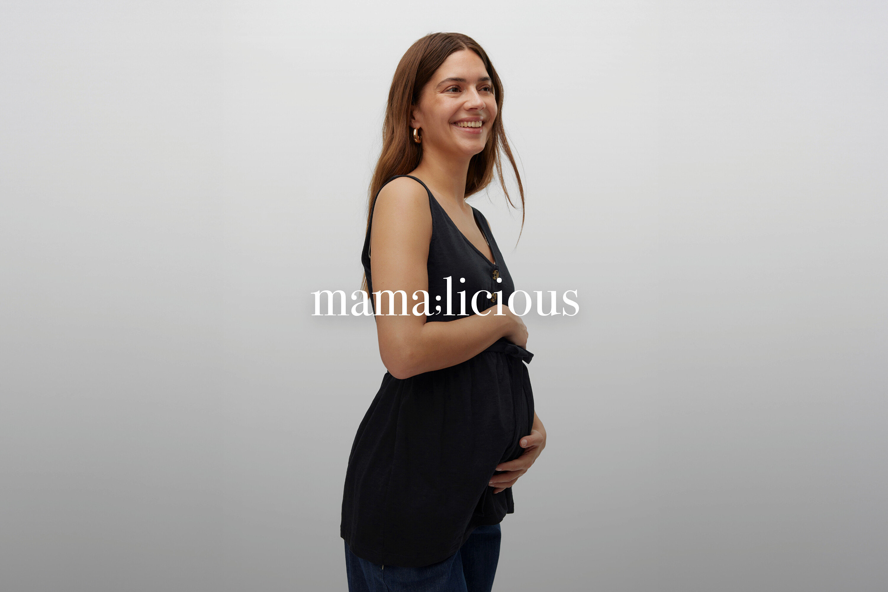 Usmievavá tehotná žena s dlhými hnedými vlasmi v čiernom tehotenskom tope bez rukávov a džínsoch.