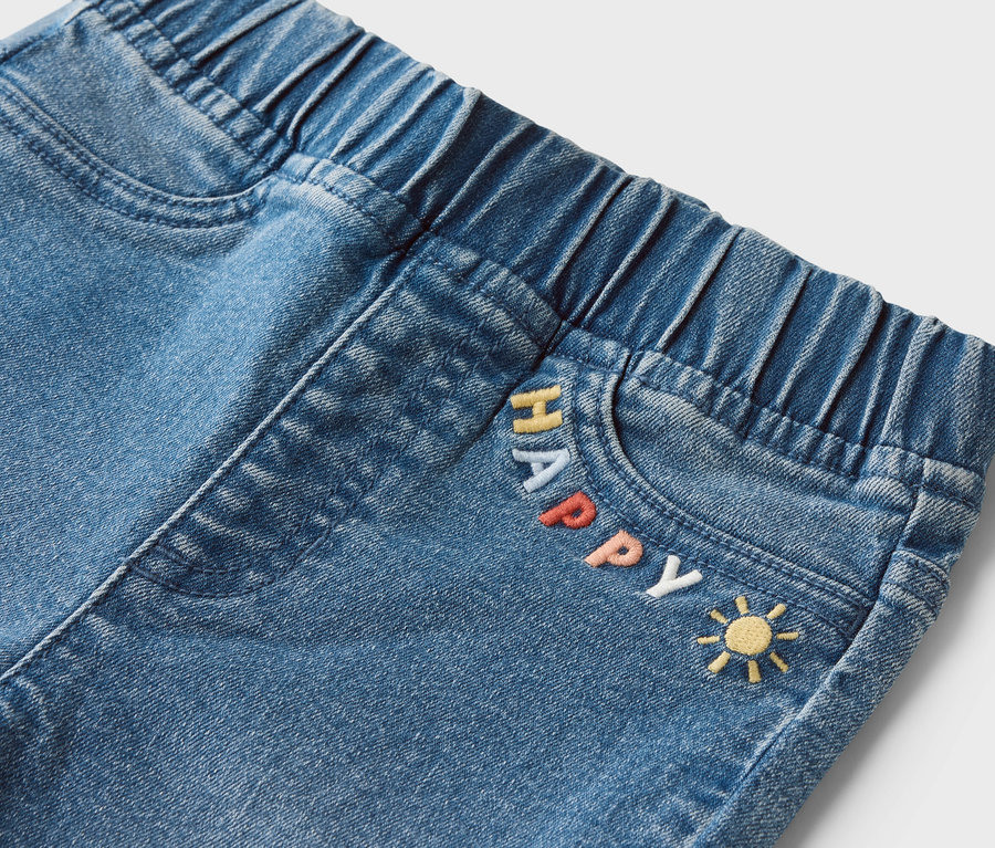 Detail džínsov s elastickým pásom a nápisom "HAPPY" a emotikonom slnka na vrecku.