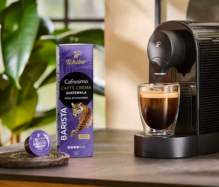 Kávovar pripravuje do sklenice kávu Cafissimo BARISTA Caffè Crema Guatemala. Vedľa stojí balenie 10 kapsúl a jedna kapsula.