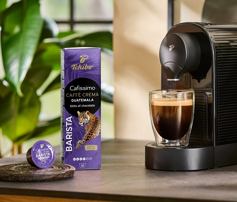 Kávovar pripravuje do sklenice kávu Cafissimo BARISTA Caffè Crema Guatemala. Vedľa stojí balenie 10 kapsúl a jedna kapsula.