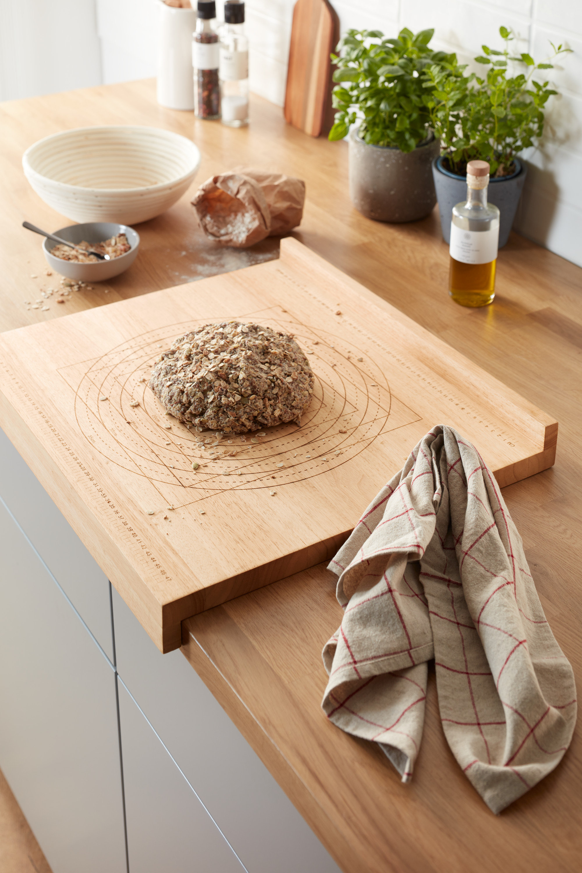 Na kuchynskej linke leží drevená pracovná doska na pečenie s bochníkom chleba. V pozadí sú bylinky v kvetináčoch a iné kuchynské potreby.