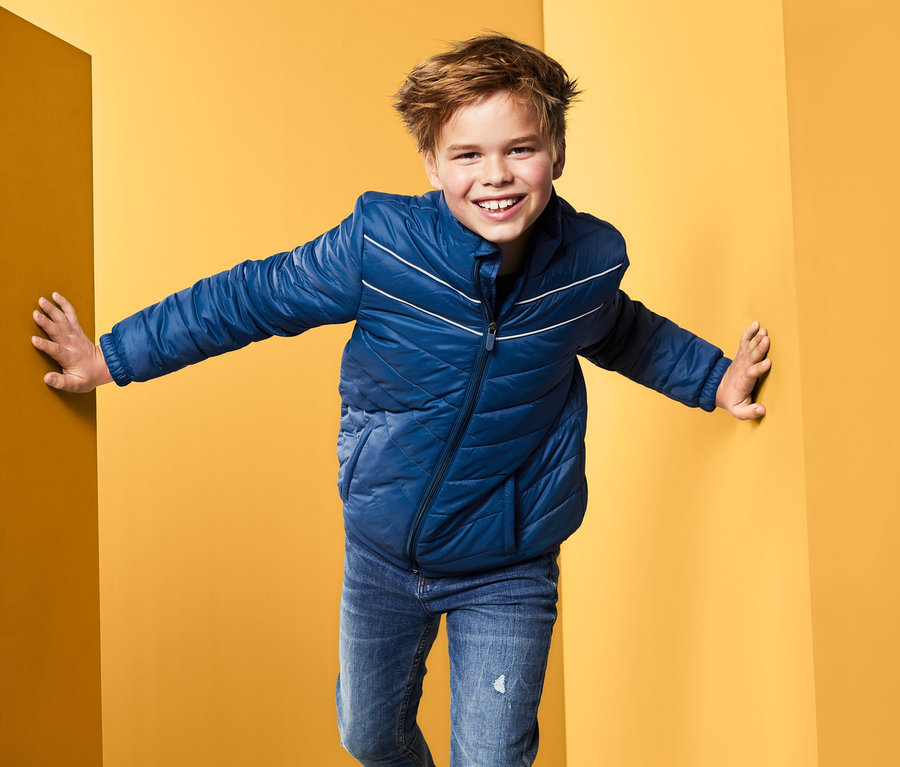 Chlapec v modrej prešívanej bunde stojí v rohu žltej miestnosti.