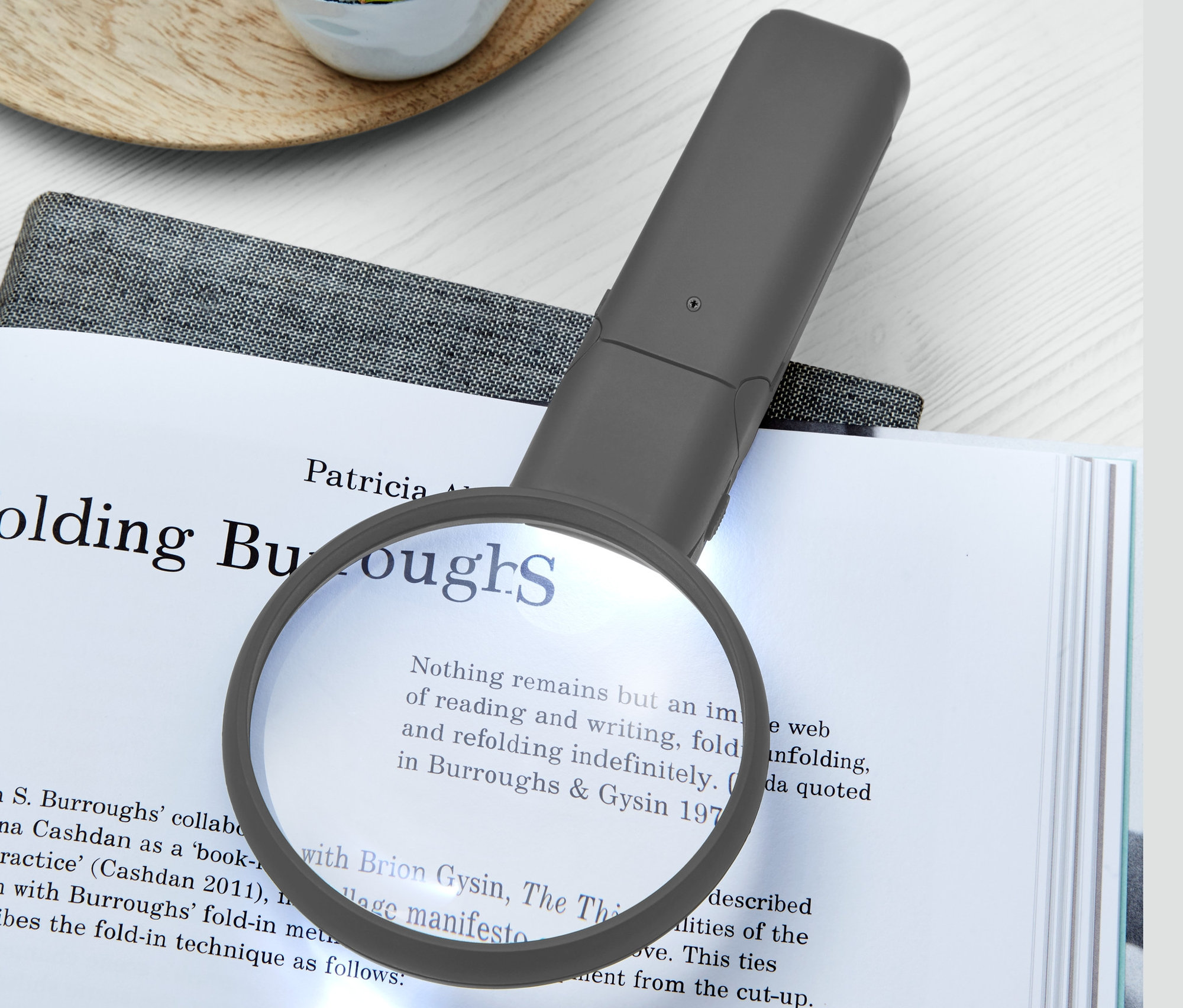 Na otvorenej knihe leží LED lupa na čítanie a vedľa nej je sukulent v bielom hrnčeku.