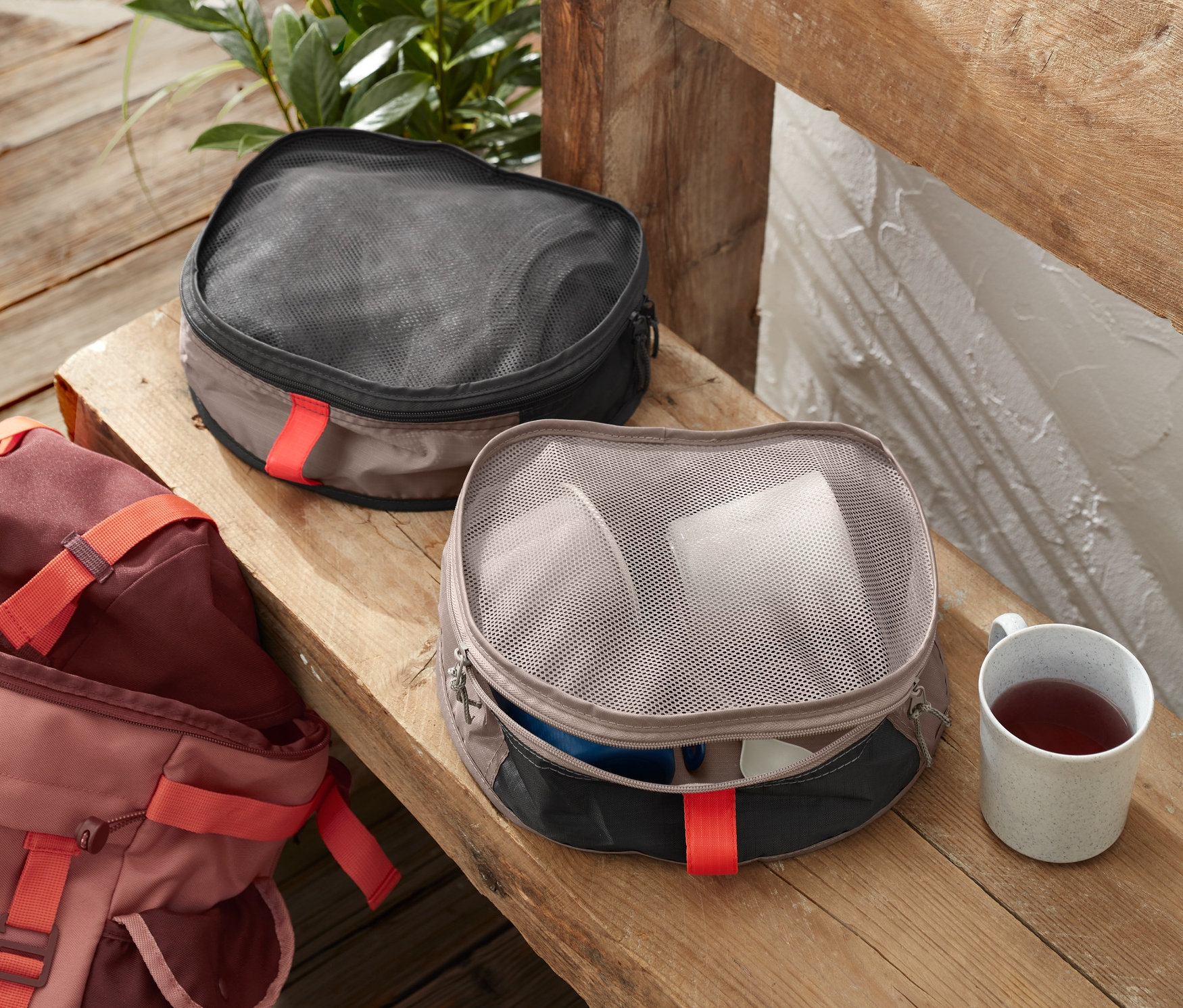Dva organizéry do ruksaku ležia vedľa seba na drevenej lavici. Jeden je tmavý a druhý svetlý. Vedľa organizérov je hrnček s čajom a ruksak.
