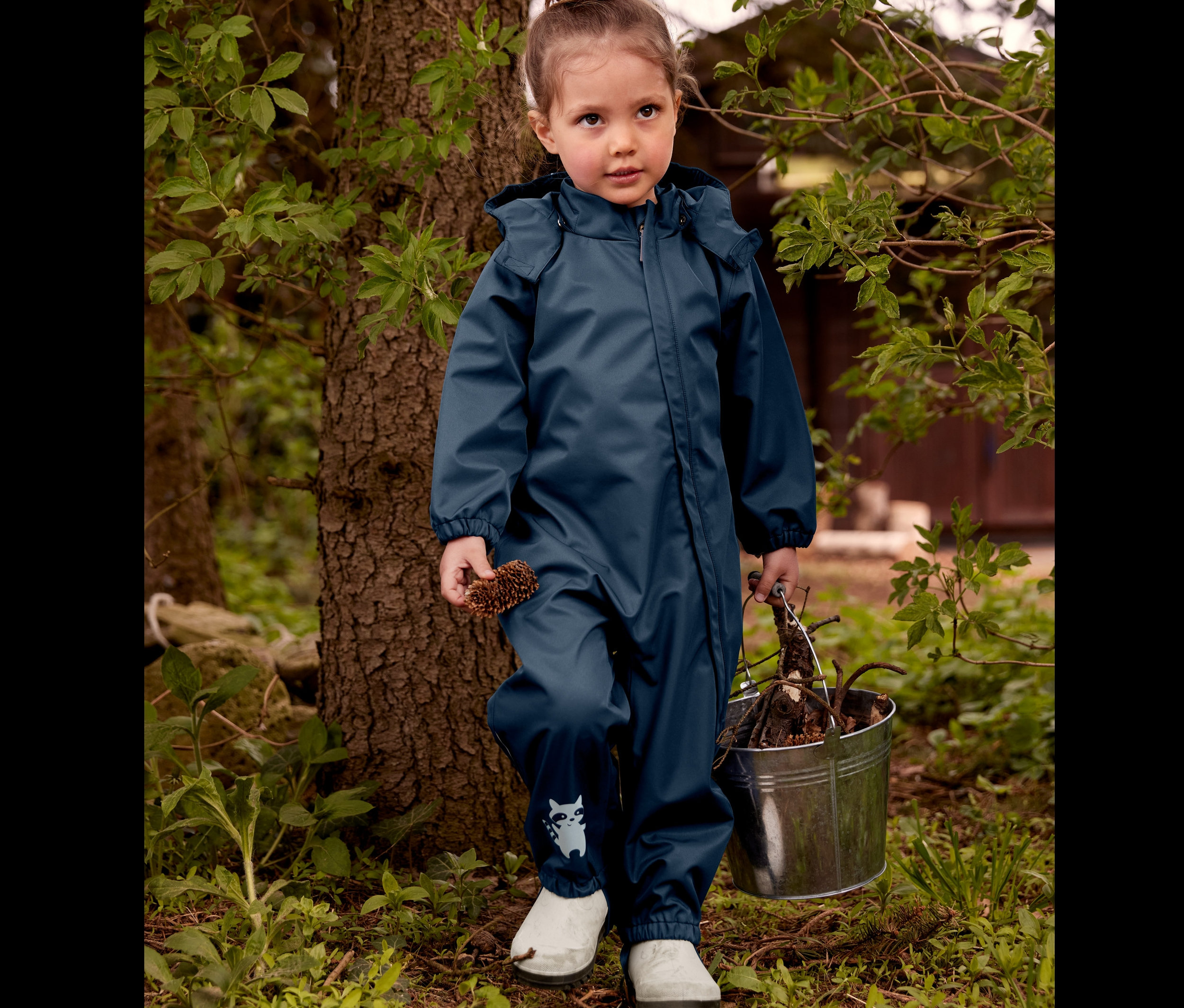 Dievčatko stojí pri strome v gumových čižmách a overale do dažďa, drží šišku a vedierko s konármi.