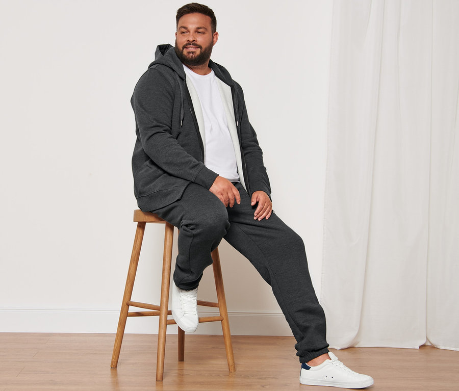 Muž sedí na drevenej stoličke a má na sebe antracitovú melírovanú mikinu s kapucňou, biele tričká s okrúhlym výstrihom (2 ks), teplákové nohavice a topánky sneaker.