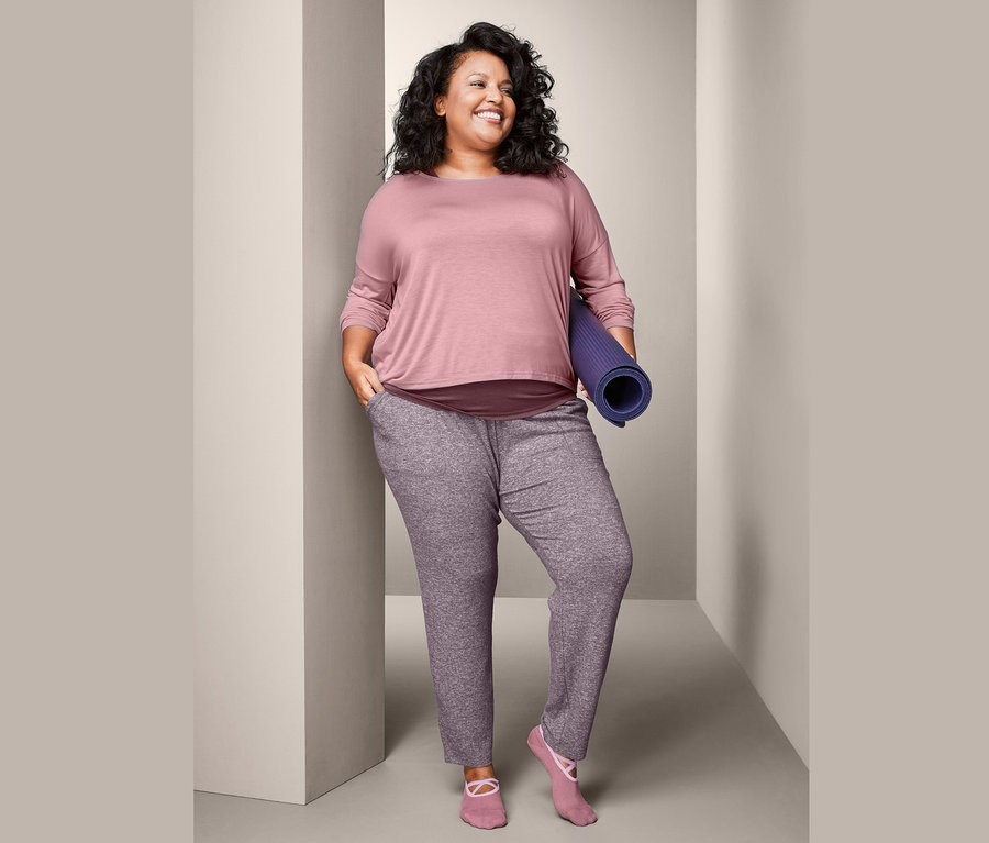 Žena drží zrolovanú podložku na cvičenie, má na sebe športové tričko 2 v 1 a pohodlné nohavice.