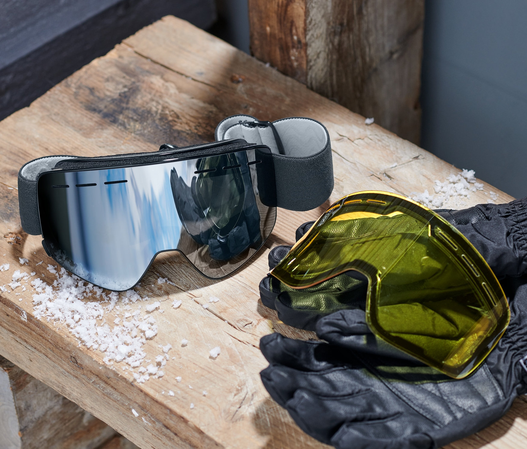Lyžiarske okuliare s magnetickými výmennými priezormi ležia na drevenej lavici vedľa žltého priezoru a čiernych rukavíc.