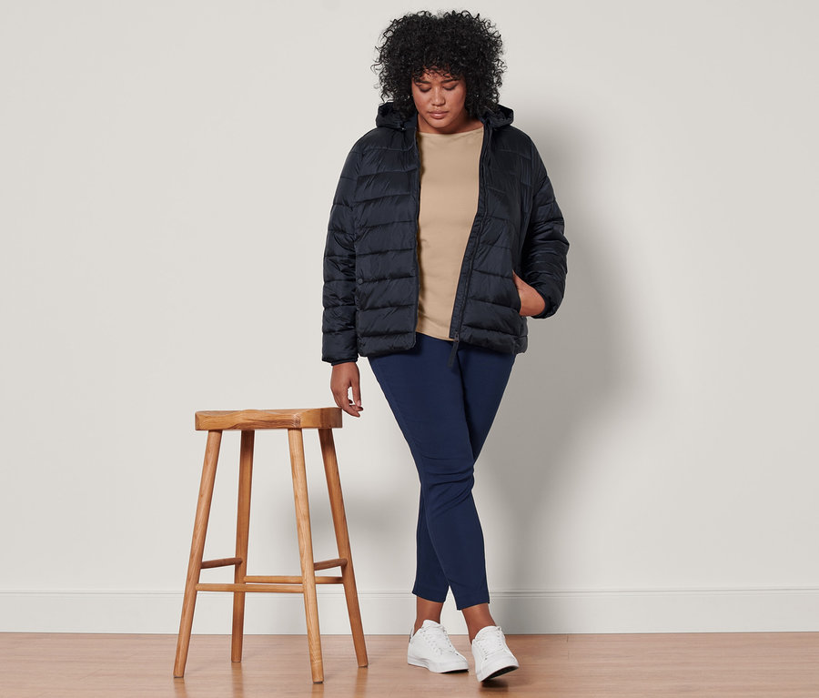Žena stojí a má na sebe tmavomodrú prešívanú bundu s kapucňou, béžové tričko s trojštvrťovými rukávmi a tmavomodré elastické nohavice.