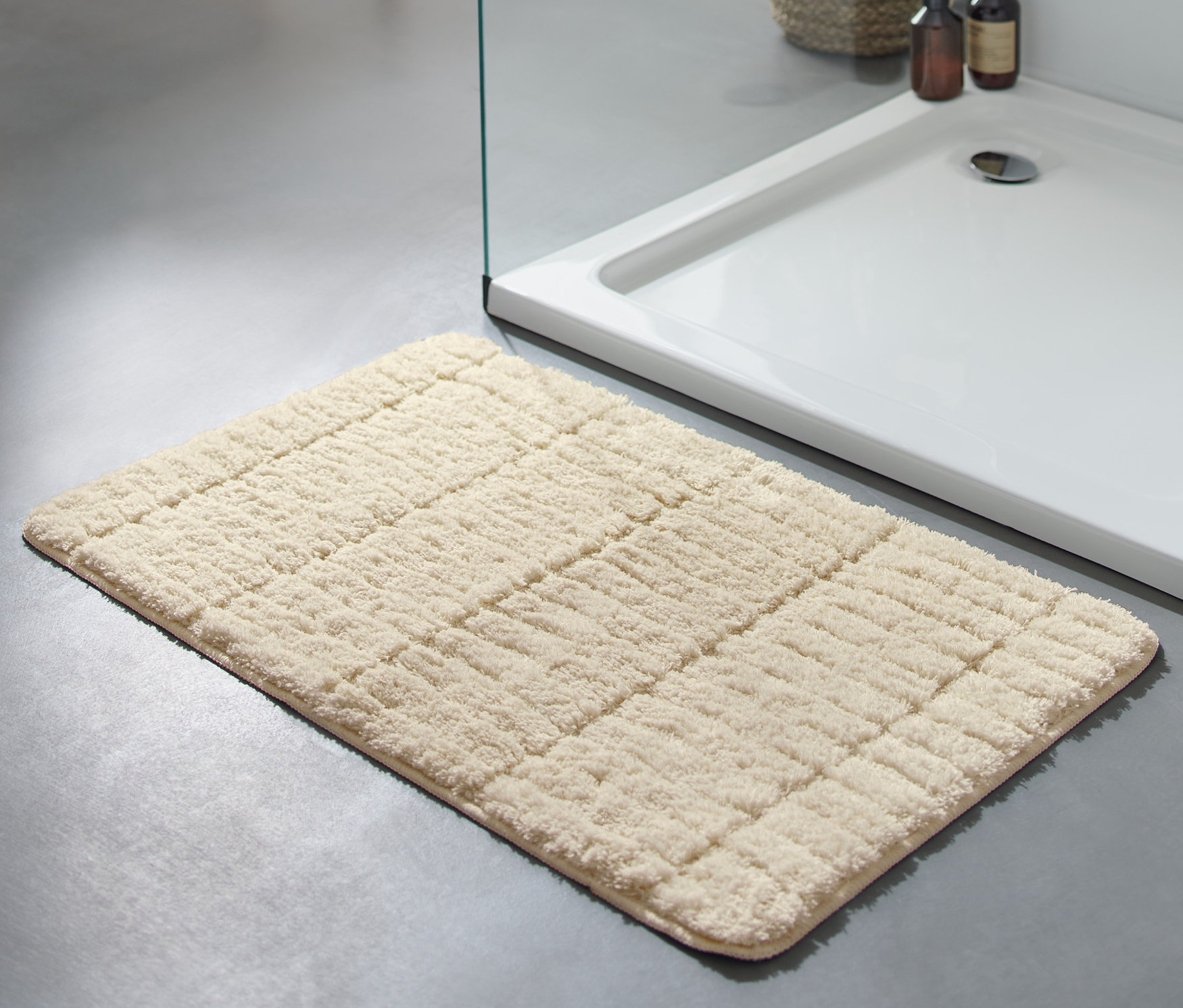 Sivobiela predložka do kúpeľne Comfort, cca 50 x 80 cm, leží na šedej podlahe vedľa sprchovacieho kúta.