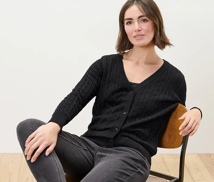 Žena sedí na stoličke a má na sebe čierny kardigán z jemnej pleteniny s vrkočovým vzorom.