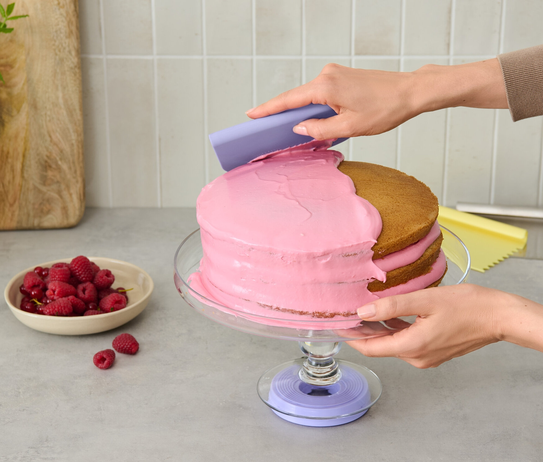Osoba nanáša ružový krém na tortu na otočnom stojane na tortu pomocou fialového nástroja.