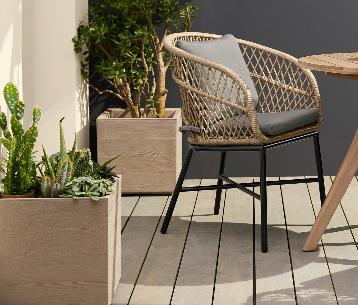 Dve jedálenské kreslá s polyratanovým výpletom stoja pri okrúhlom záhradnom stolíku. Nad stolom visí závesné solárne svietidlo, filamentové. Vľavo stojí kvetináč na kolieskach.