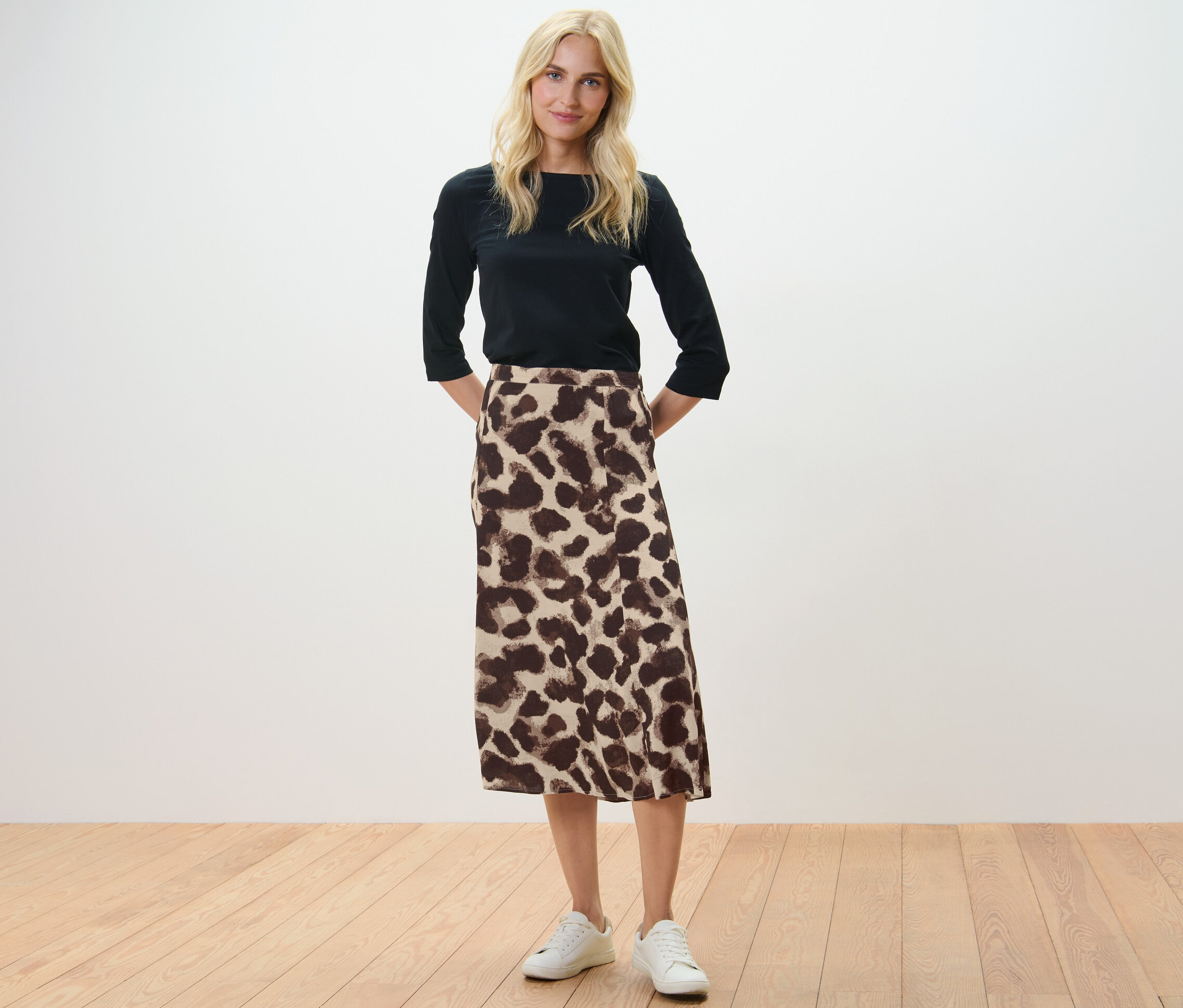 Blondínka stojí a má ruky za chrbtom. Oblečená je do čiernej blúzky a midi sukne s potlačou.