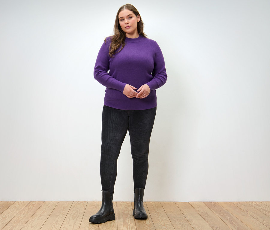Žena pózuje v pulóvri z jemného úpletu a menčestrových legínach.