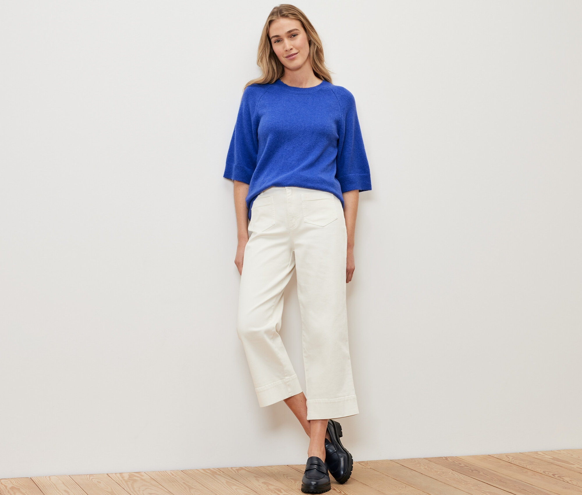 Žena stojaca pred bielou stenou má na sebe pletený pulóver kobaltovomodrej farby, biele nohavice a čierne topánky.