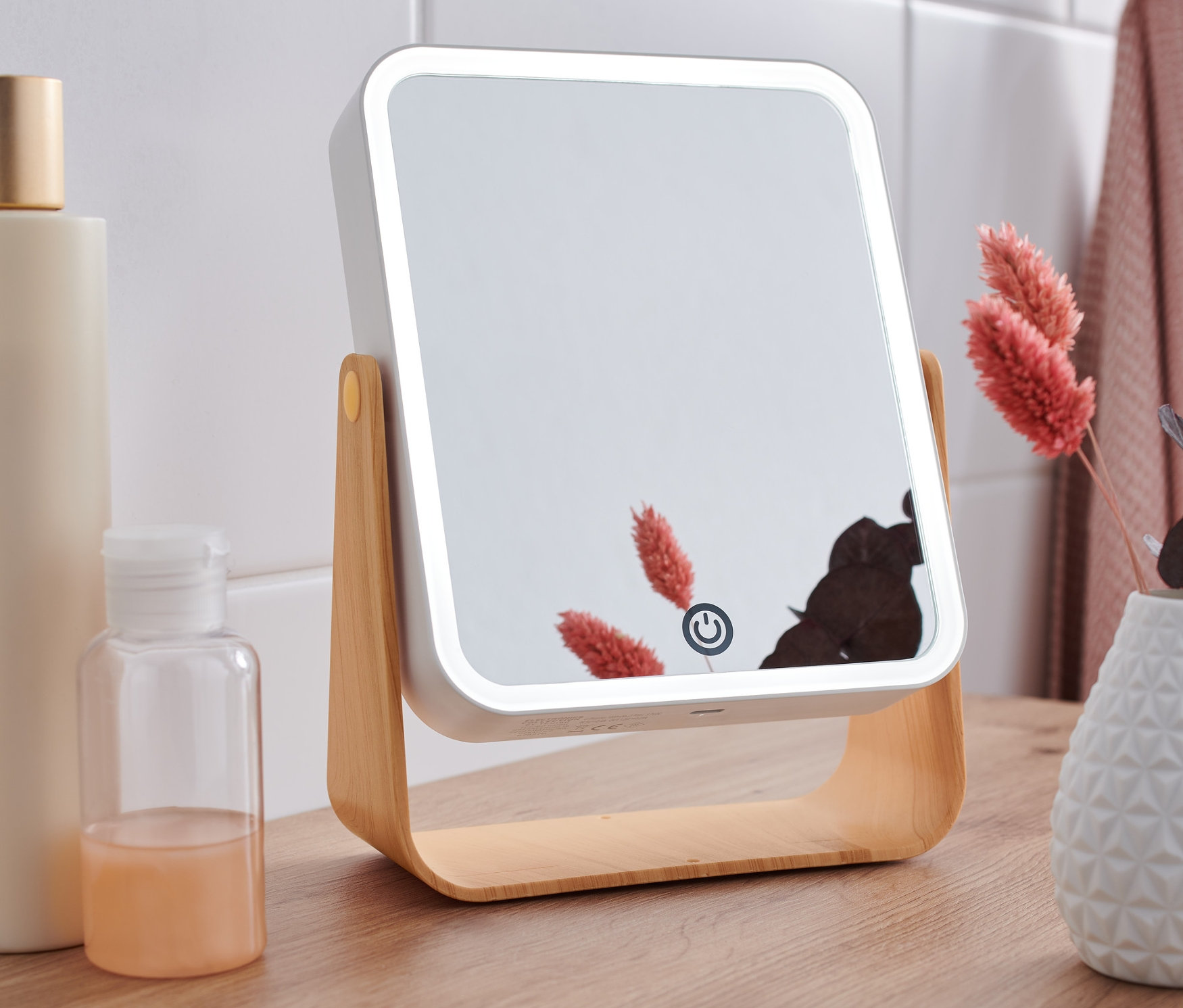 Kozmetické zrkadlo s LED osvetlením stojí na drevenom stole vedľa fľaštičiek a vázy s kvetinami.