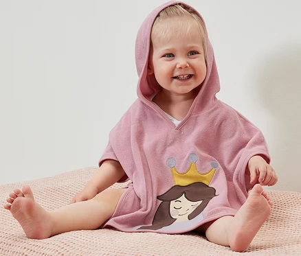 Usmievajúce sa dieťa sedí na ružovom povrchu a má na sebe froté pončo s kapucňou s motívom princeznej.