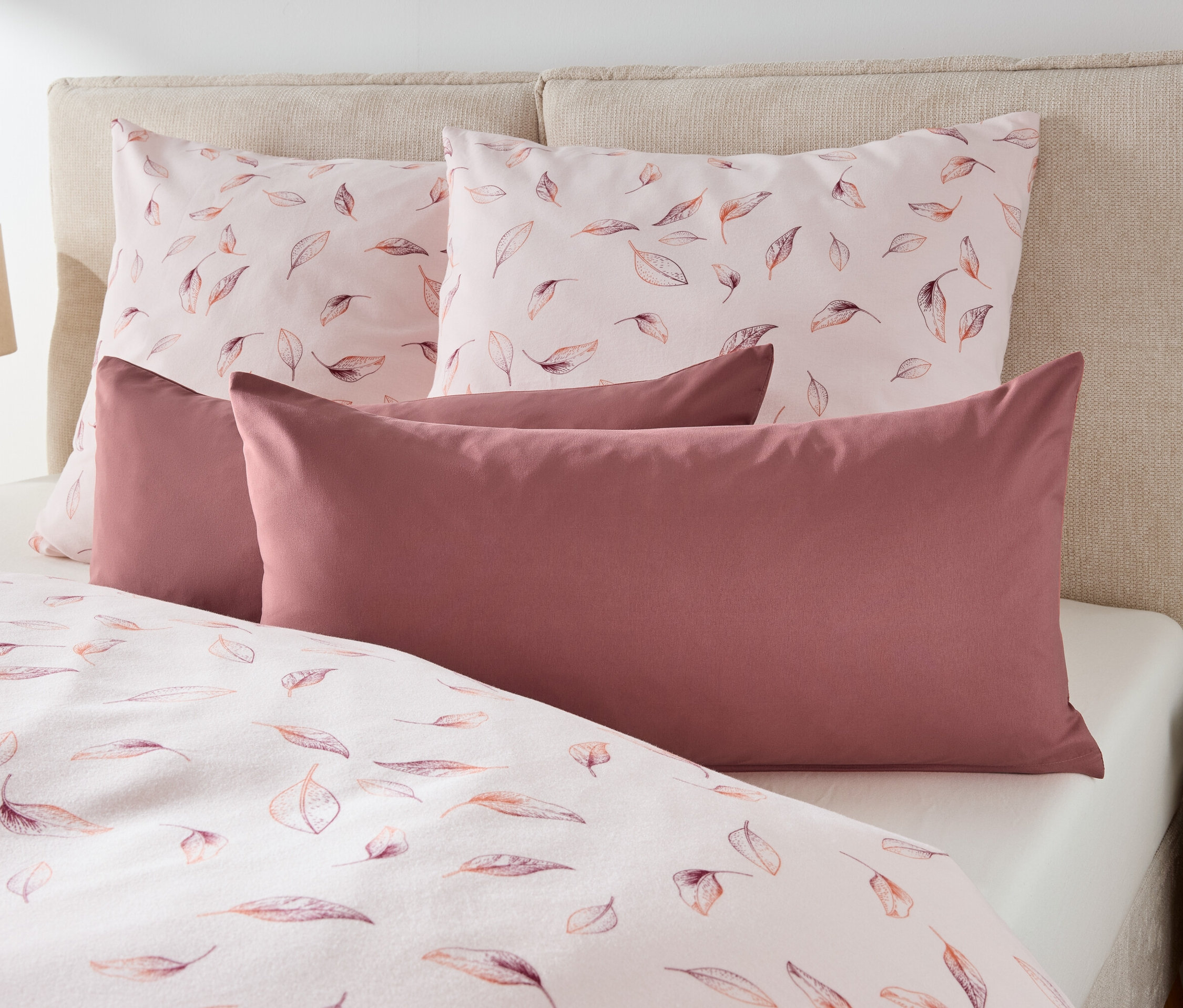Na posteli sú dva vankúše s potlačou listov a dva bordové vankúše.