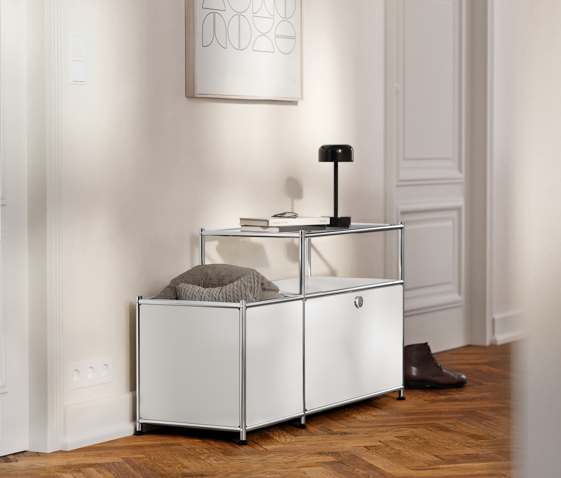 V chodbe stojí biely kovový lowboard »CN3« s výklopnými dvierkami a sklenenou doskou, na ktorom leží kniha a lampa.