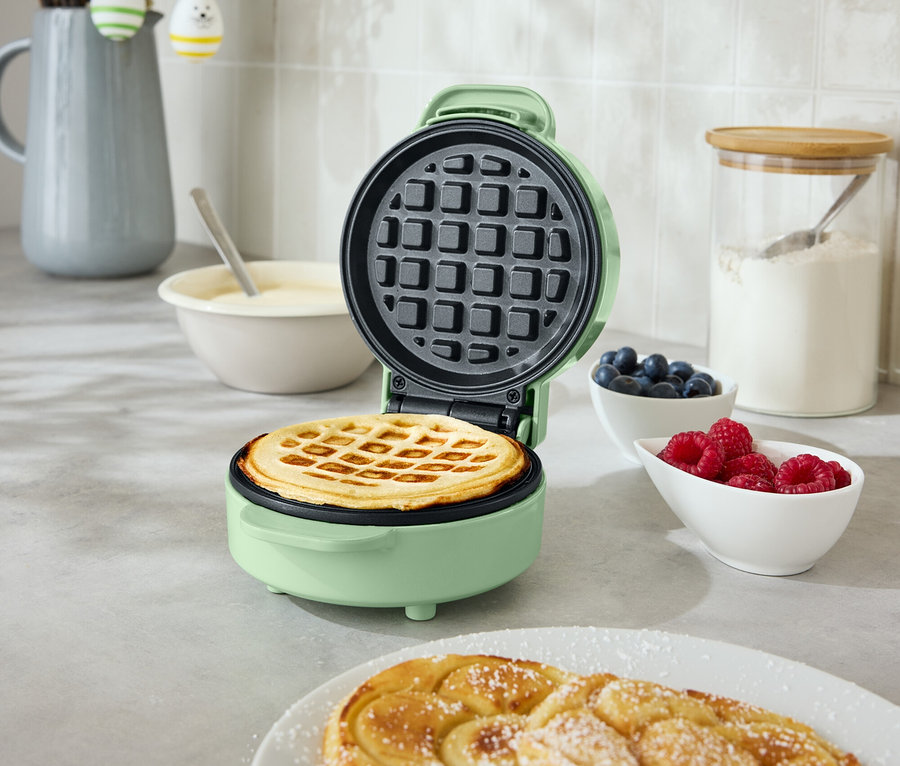 Otvorený, okrúhly, mätovo zelený vaflovač s vafľou vnútri stojí na kuchynskej doske. Vedľa sú dva špirálovité pečené výrobky na tanieri, miska s čučoriedkami a miska s malinami.