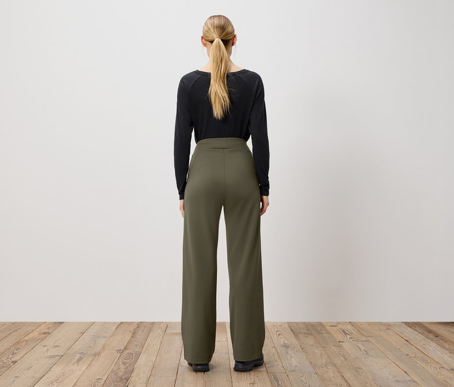 Žena stojaca chrbtom k fotoaparátu má na sebe čierne tričko s dlhým rukávom a olivovozelené nohavice.