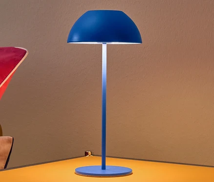 Modrá stolová LED lampa s akumulátorom na žltom nočnom stolíku.