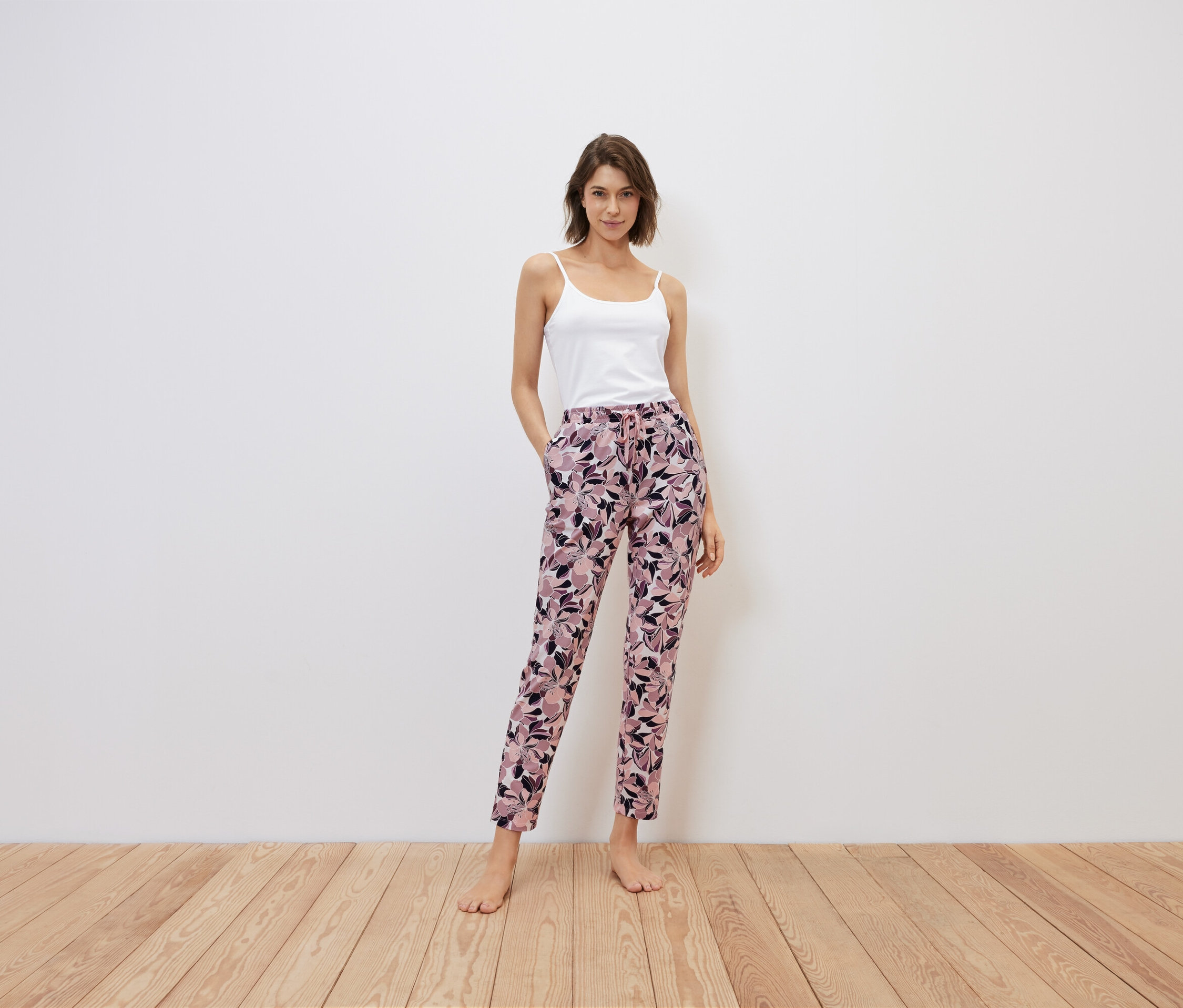 Žena stojí a má ruky vo vreckách kvetinových nohavíc, biely top.