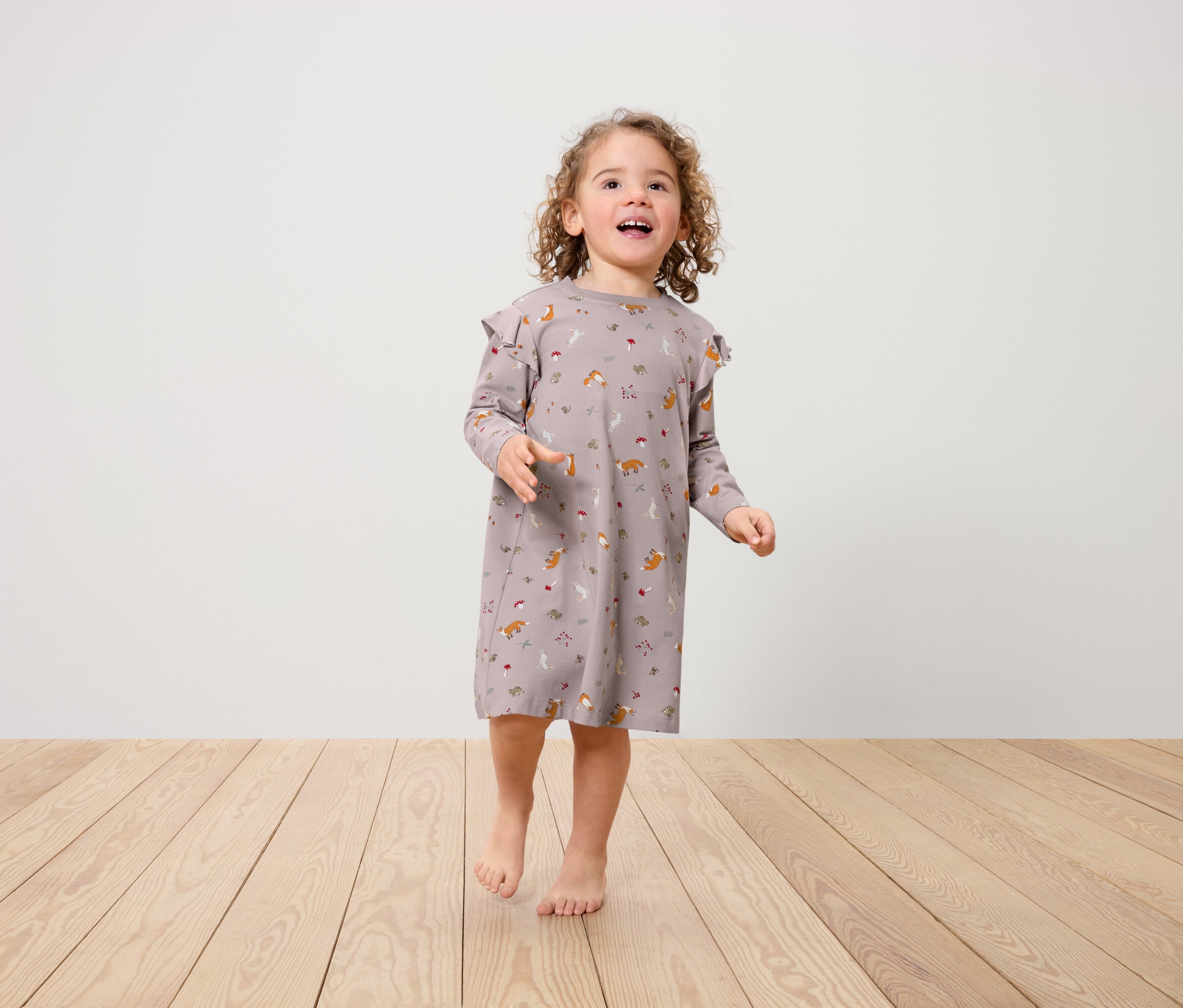 Dievčatko v šatách s potlačou líšok a volánikmi na ramenách stojí na drevenej podlahe.