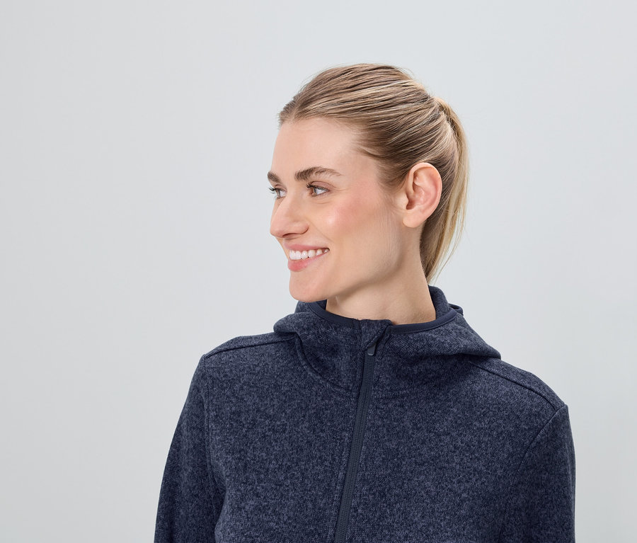 Profil usmievajúcej sa ženy s blond vlasmi v modrej mikine s kapucňou na zips.