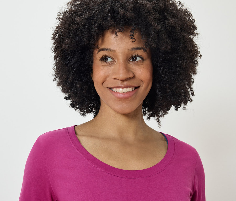 Portrét usmievajúcej sa ženy s afro účesom v ružovom tričku, pozerajúcej sa doľava.