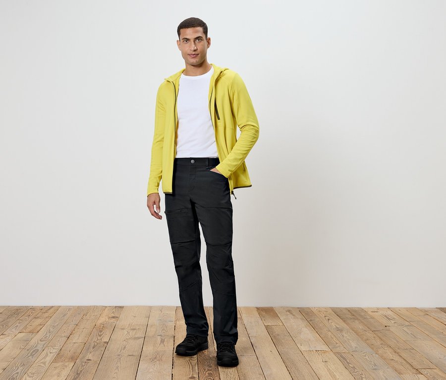 Muž stojí na drevenej podlahe. Má na sebe čierne nohavice, biele tričko a žltú mikinu.