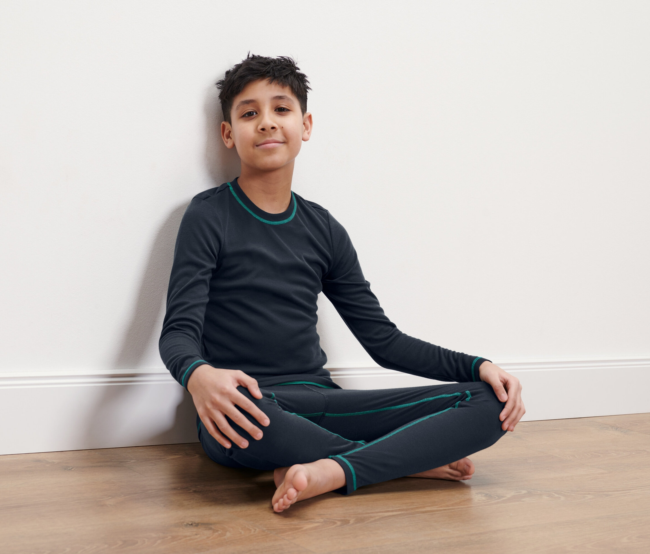 Chlapec sedí so skríženými nohami na podlahe a opiera sa o bielu stenu. Má na sebe tmavomodrú termobielizeň.