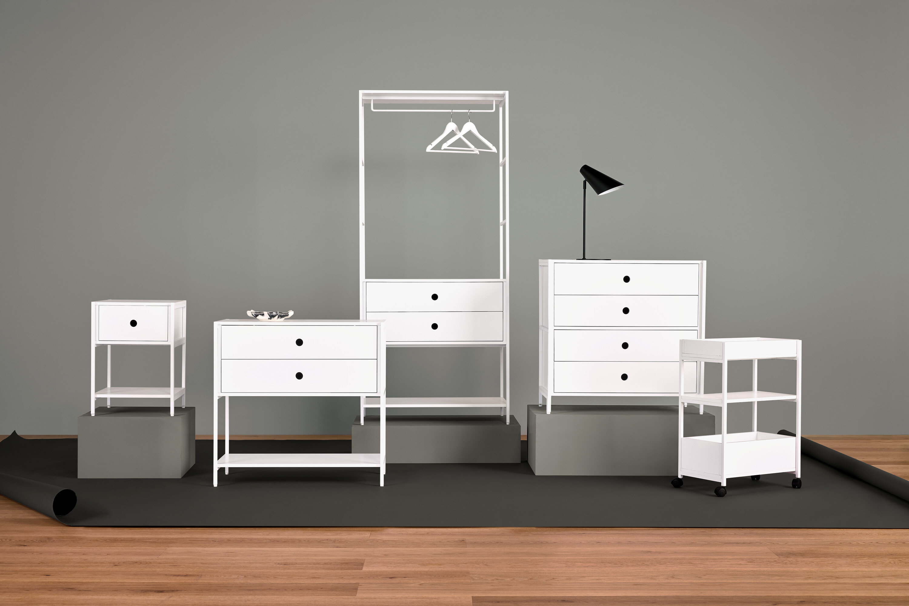 Usporiadanie bieleho nábytku s nočným stolíkom, komodou so zásuvkami, vešiakom na šaty, lampou a policami.