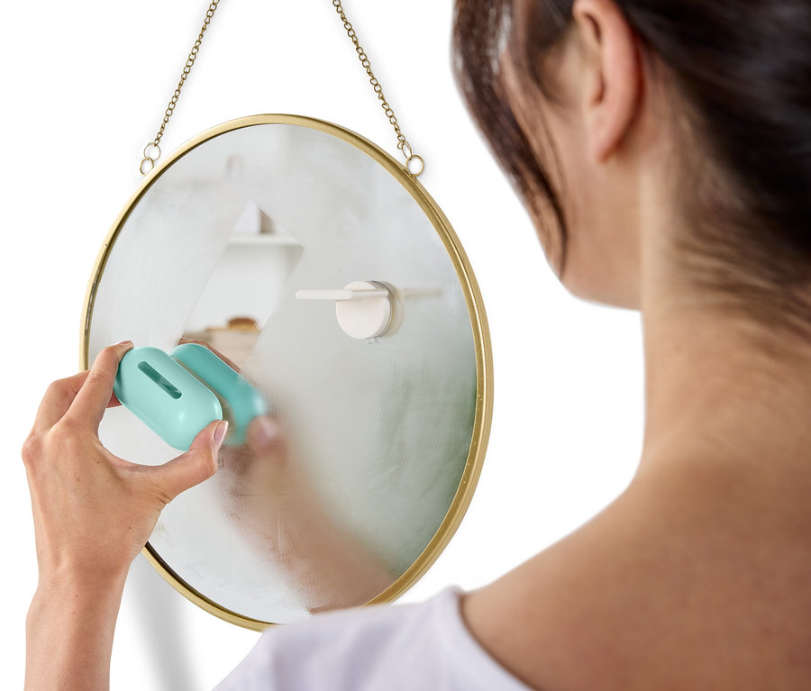 Žena čistí okrúhle zrkadlo zavesené na stene pomocou modrého čističa.