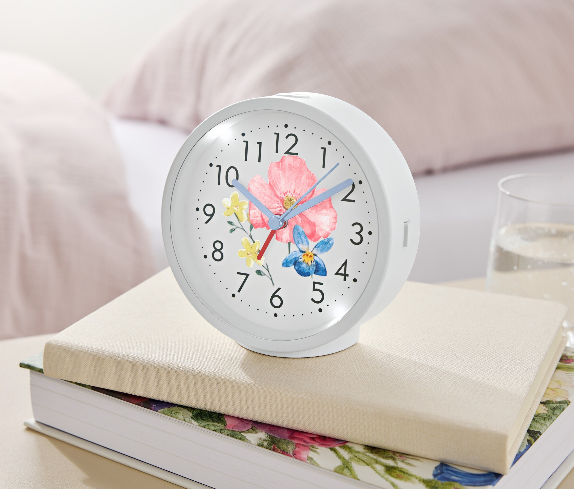 Biely budík s kvetinovým motívom leží na knihách vedľa pohára vody.