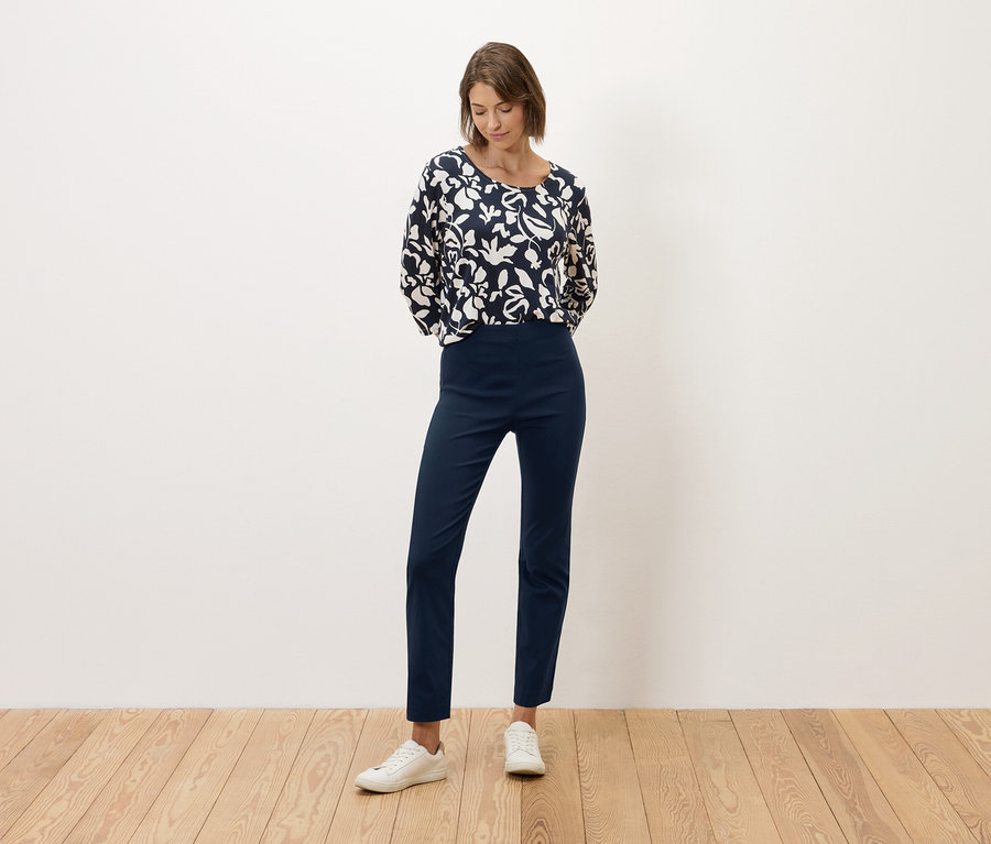 Žena stojí pred bielou stenou, oblečená v tmavomodrých nohaviciach a blúzke s čierno-bielou potlačou.