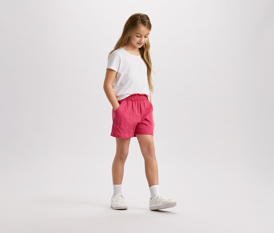 Dievča s blond vlasmi, v bielom tričku a ružových šortkách stojí a pozerá sa dole.