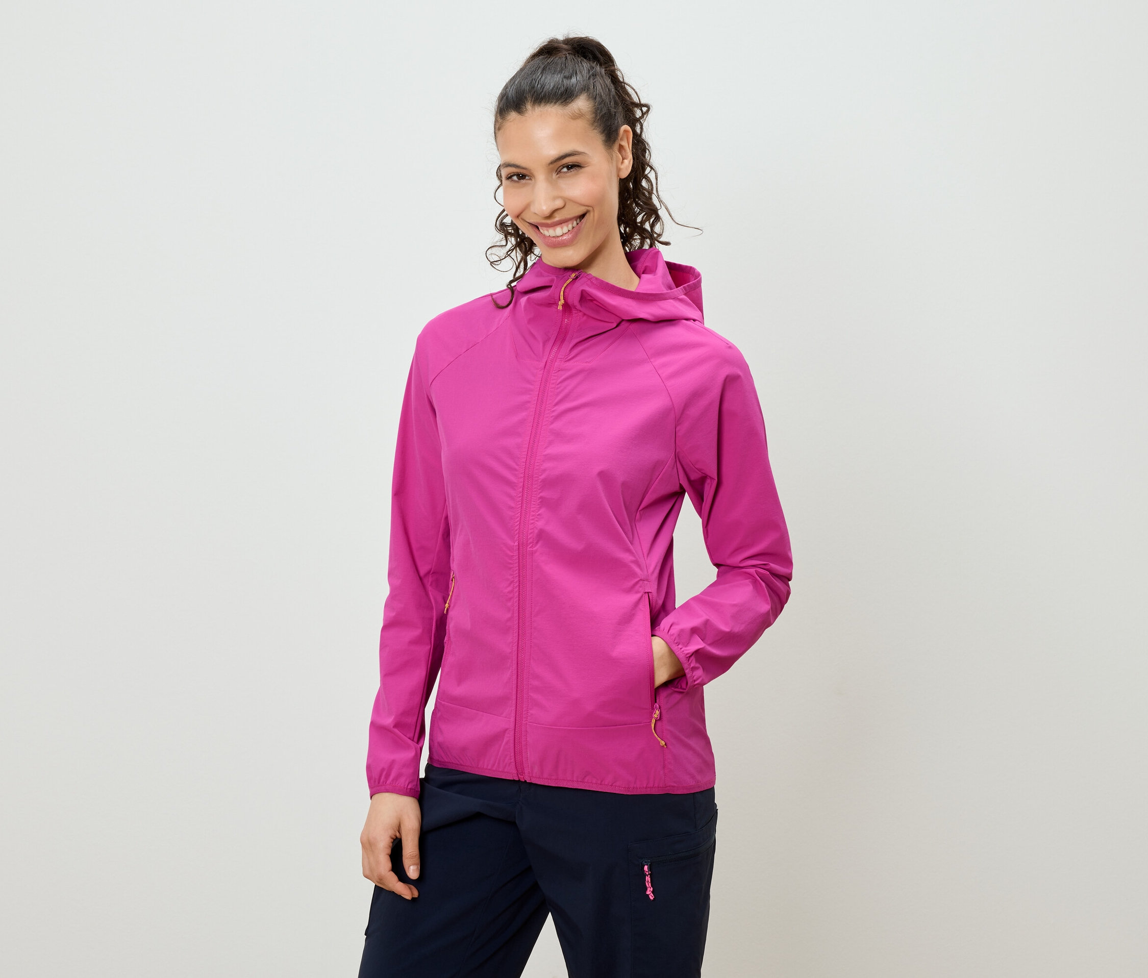 Usmievavá žena s vlasmi zopnutými do chvosta má na sebe ružovú bundu s kapucňou a tmavomodré nohavice.