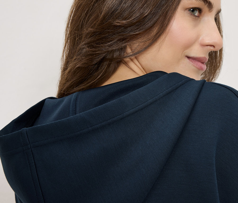 Žena má na sebe tmavomodrú mikinu s kapucňou, pozerá cez plece.