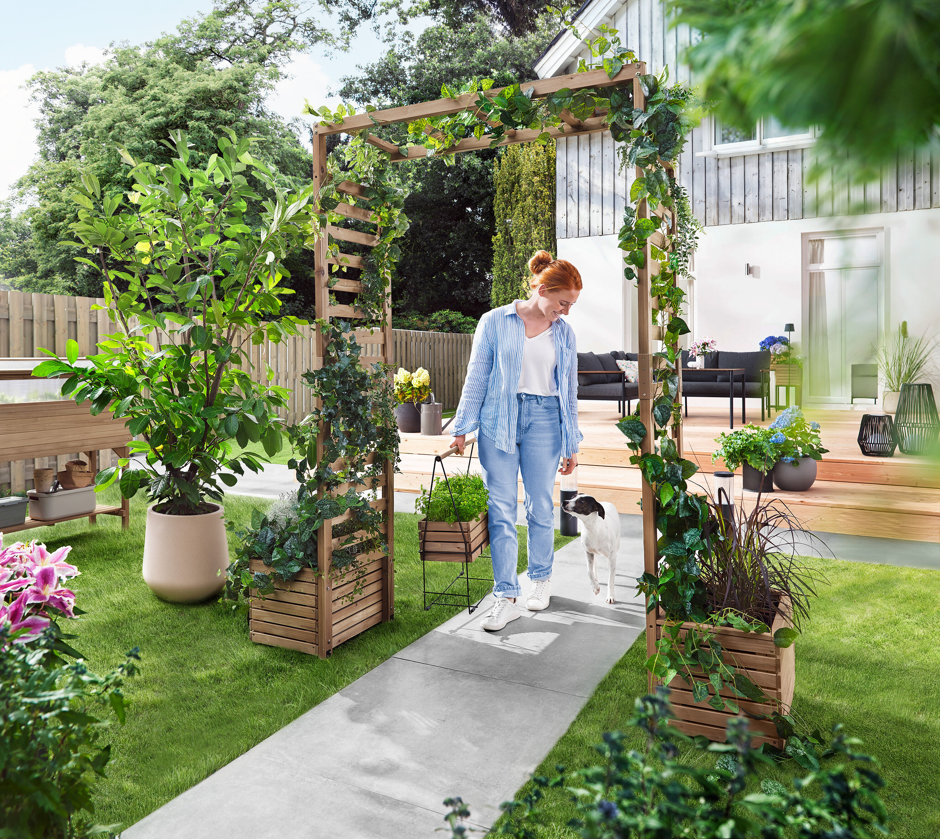 Žena prechádza so psom cez záhradný oblúk porastený rastlinami. Nesie kvetináč.