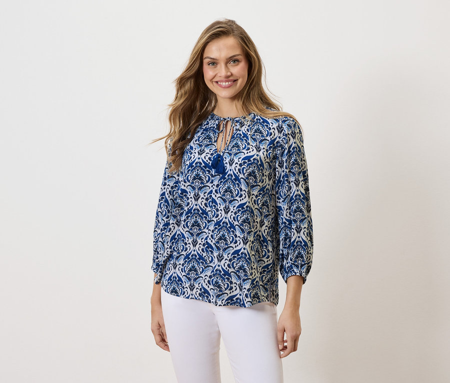 Žena s dlhými blond vlasmi sa usmieva a má na sebe modro-biely vzorovaný top a biele nohavice.