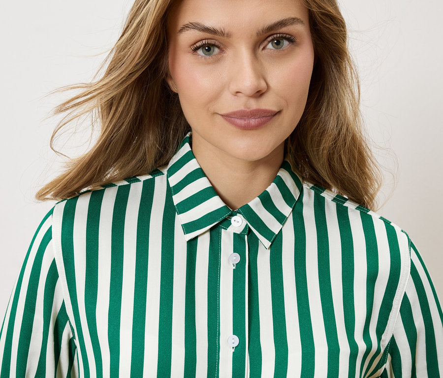 Portrét ženy s blond, vlnitými vlasmi, oblečenej v pruhovanej košeli smaragdovo zelenej a bielej farby.