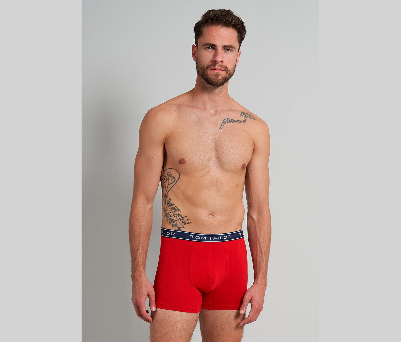 Muž s tetovaniami má na sebe červené boxerky Tom Tailor na sivom pozadí.
