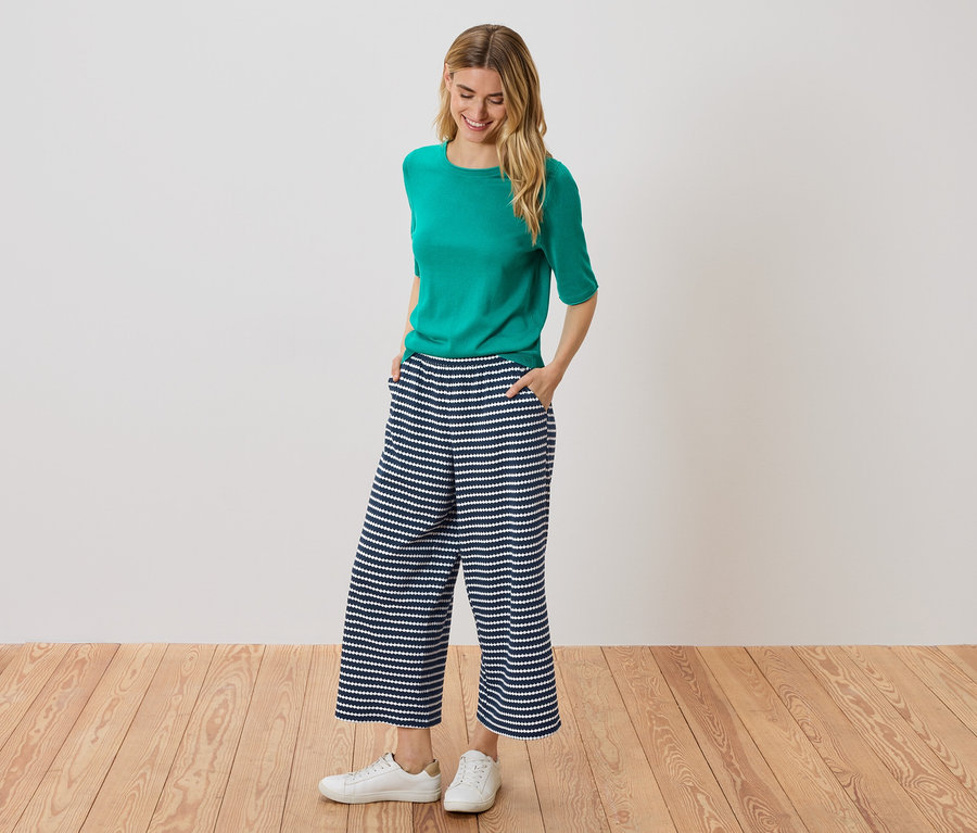 Žena stojí s rukami vo vreckách v modro-bielych pruhovaných culottes, má na sebe zelený top a biele tenisky.