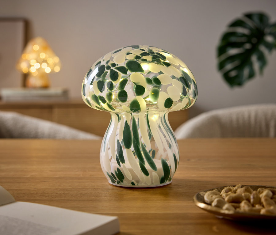 Lampa v tvare huby so zeleno-bielym vzorom a svetielkami stojí na drevenom stole. Vedľa nej je otvorená kniha a malá miska s kešu orieškami. V pozadí je vidieť ďalšiu lampu v tvare huby a rastlinu.